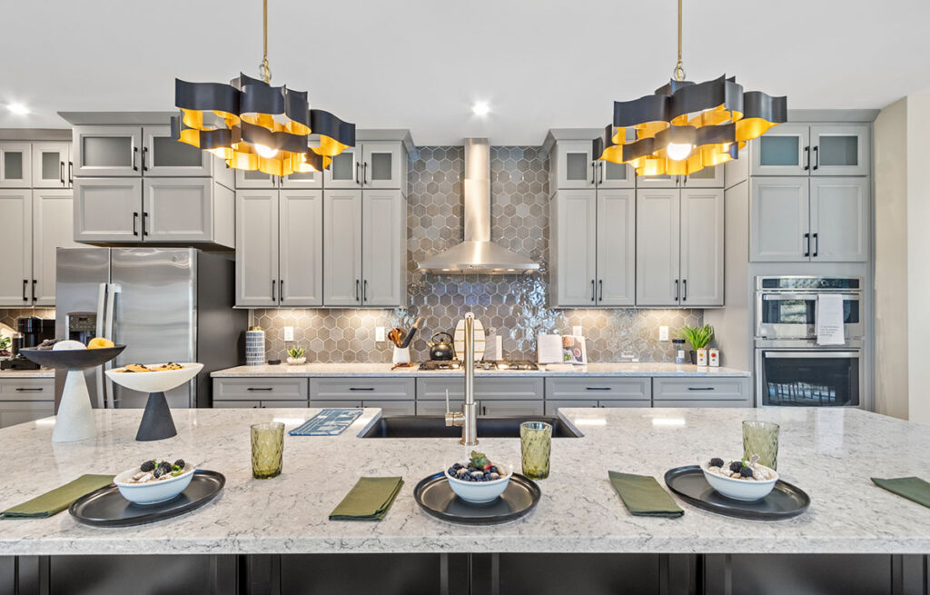 Modern kitchen inside a Fischer Homes house featuring a long gray island with marble countertop set for dinner, gray cabinets with stainless appliances, and modern gold pendant lights.