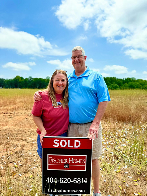 Smiling couple stands together in a grassy field next to a red “SOLD” sign for Fischer Homes after purchasing a new home site