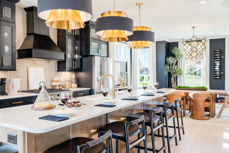 Modern kitchen interior of a Fischer Homes model featuring a long island with bar stools, dark cabinetry, gold pendant lights, and decorative table settings.