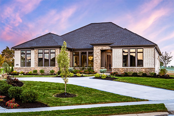 Single-story modern ranch-style house built by Fischer Homes featuring a dark roof, large front windows, and a stone and brick facade with a landscaped yard under a colorful sunset