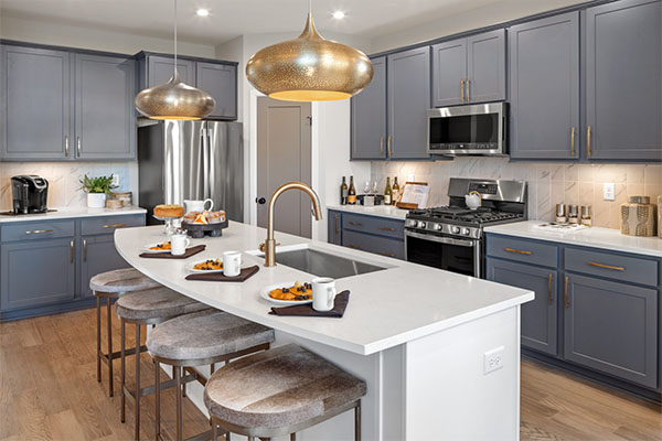Modern kitchen inside a Fischer Homes house featuring a large white island with gray cushioned barstools, blue cabinetry, gold pendant lights, and stainless steel appliances