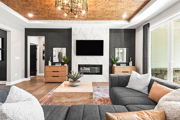 Modern living room inside a Fischer Homes house featuring a gray sectional sofa, fireplace with mounted TV, wooden ceiling with chandelier, and decorative plants.