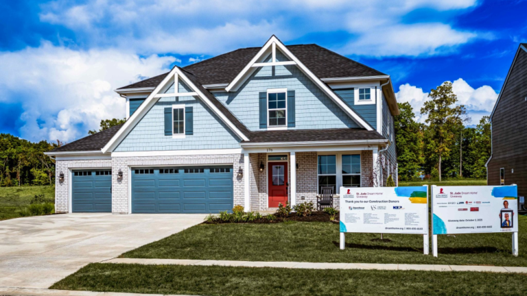 Two-story blue house built by Fischer Homes with white trim, double garage and a St. Jude sign in the front yard under a blue sky.