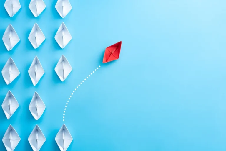 Illustration of a red paper boat veering away from a formation of white paper boats on a blue background, symbolizing innovation or leadership and opportunity for growth.