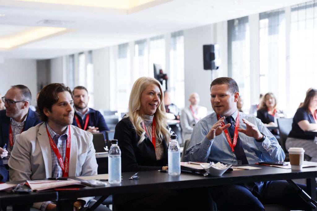 Group of professionals engaged in discussion at a conference table wearing red lanyards during a meeting in a bright conference hall.