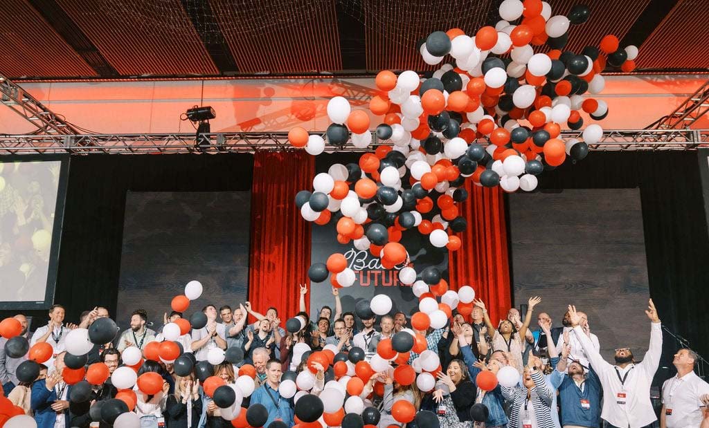 Fischer Group associates celebrate at an indoor event, releasing red, white, and black balloons under a stage with banners.